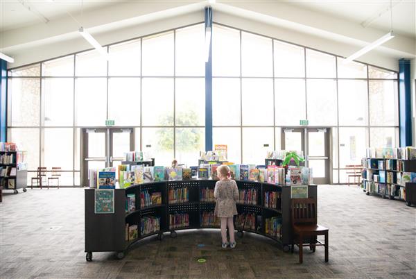 Black and white photo of student selecting a book with tall library windows offering filtered sunlight